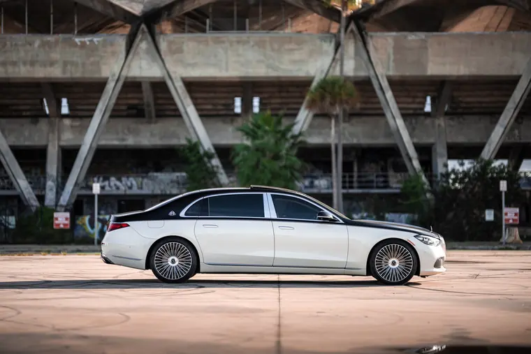 Two-tone Mercedes-Maybach S580 rental in side profile, cream over black with the Maybach monogram on the rear pillar, parked on open concrete in front of a graffitied grandstand and palm trees in Miami