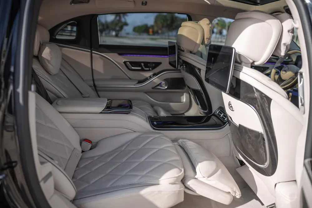 Rear cabin of the Mercedes-Maybach S580 rental with the right-hand seat reclined and its calf rest raised, white diamond-quilted leather, loose pillows and a screen on the seatback